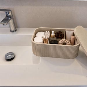 Könnte beinhalten: Eine beige, rechteckige Aufbewahrungsbox mit gerippter Oberfläche und passendem Deckel, die mehrere Innenfächer enthält, gefüllt mit Wattepads, Wattestäbchen, einer kleinen Glastropfflasche, Haarspangen, einem beigen Haargummi und Kosmetiktuben.