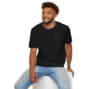 May include: Black t-shirt with the initials "B.B." in white on the upper left chest. The shirt is short-sleeved and worn with blue jeans. The person is sitting on a white surface.