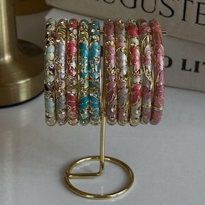 May include: A collection of ten thin metal bangles displayed on a gold-toned stand. Each bangle features a repeating floral pattern with enamel fill in shades of blue, pink, and white, accented with small clear crystals set into the metal frame.