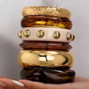 May include: A stack of six bangles featuring a variety of textures and finishes. The set includes a hammered gold-tone bangle, a translucent amber resin bangle, a cream-colored bangle with gold studs, a dark brown marbled resin bangle, a smooth polished gold-tone bangle, and a dark brown tortoiseshell-patterned resin bangle.