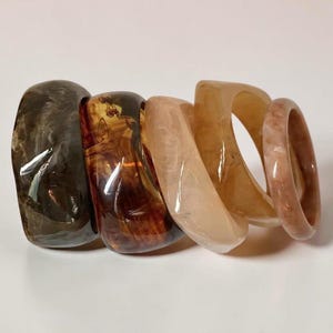 Puede incluir: Un conjunto de cinco brazaletes de resina lisa y pulida dispuestos en fila. Las pulseras presentan un degradado de colores que va desde el gris carbón oscuro hasta el ámbar profundo, seguido de un beige claro, un bronceado translúcido y un rosa viejo jaspeado.