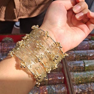 May include: A stack of clear, translucent bangles featuring embedded gold-toned glitter accents and small, dangling gold-coloured metal charms attached to the outer edges of the bracelets, creating a layered and reflective appearance on the wrist.