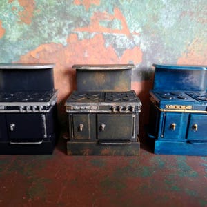 Peut inclure: Trois cuisinières miniatures disposées côte à côte. La cuisinière de gauche est noir mat avec des accents argentés. Celle du centre présente une finition sombre vieillie et usée. La cuisinière de droite est peinte en bleu vif avec des détails dorés.