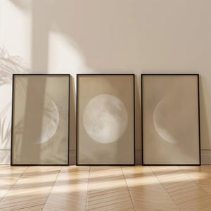 Könnte beinhalten: Ein Set aus drei rechteckigen Wandbildern in schwarzen Rahmen, die jeweils eine beige Darstellung des Mondes in verschiedenen Phasen zeigen, darunter ein zunehmender Mond links, ein Vollmond in der Mitte und ein abnehmender Mond rechts.