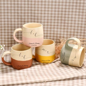 Puede incluir: Cuatro tazas de cerámica con asas, cada una con un diseño de dos tonos. Las tazas son de color crema con secciones rosas, marrones, amarillas y verdes. Cada taza tiene iniciales y una fecha impresa. Las tazas están sobre una superficie a cuadros.