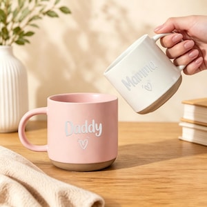 Puede incluir: Una taza de cerámica rosa con la palabra Daddy y un corazón impresos en blanco, y una taza de cerámica blanca con la palabra Mamma y un corazón impresos en blanco. Ambas tazas tienen un acabado mate liso y una base natural sin esmaltar.