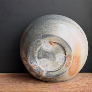 May include: A handmade ceramic bowl with a rounded shape, featuring a blend of gray, white, and orange hues. The bowl's underside is visible, showcasing a textured surface and a circular base. The bowl is placed on a wooden surface.