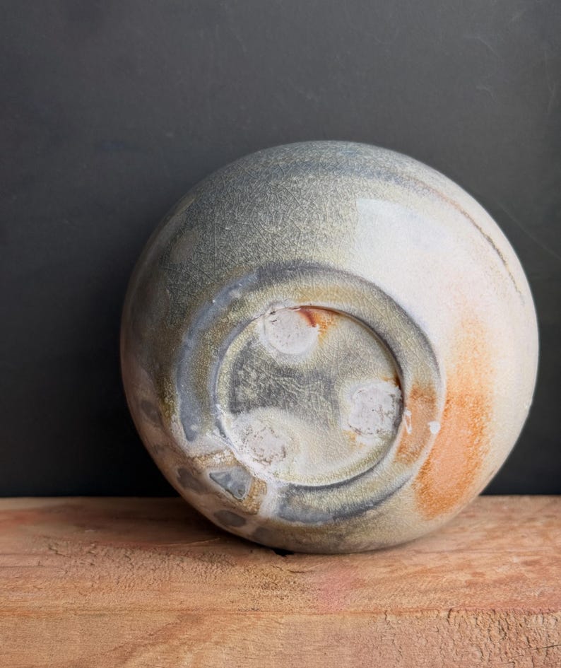 May include: A handmade ceramic bowl with a rounded shape, featuring a blend of gray, white, and orange hues. The bowl's underside is visible, showcasing a textured surface and a circular base. The bowl is placed on a wooden surface.
