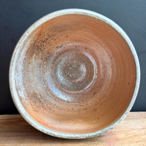 May include: A handmade ceramic bowl with a rustic aesthetic. The bowl features a warm, earthy color palette with shades of orange, brown, and white. The interior has a textured surface, and the rim is finished with a white glaze. The bowl sits on a wooden surface.
