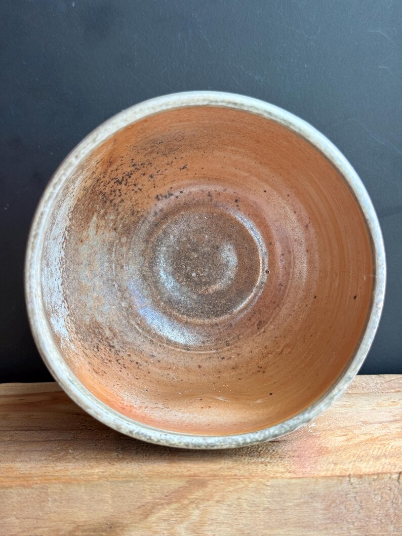 May include: A handmade ceramic bowl with a rustic aesthetic. The bowl features a warm, earthy color palette with shades of orange, brown, and white. The interior has a textured surface, and the rim is finished with a white glaze. The bowl sits on a wooden surface.