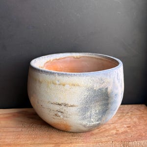 May include: A handmade ceramic bowl with a rounded shape. The bowl features a textured, off-white exterior with patches of gray and orange. The interior has an orange hue. The bowl sits on a wooden surface.