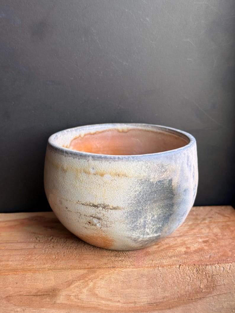 May include: A handmade ceramic bowl with a rounded shape. The bowl features a textured, off-white exterior with patches of gray and orange. The interior has an orange hue. The bowl sits on a wooden surface.