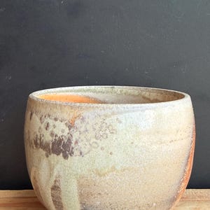 May include: A handcrafted ceramic bowl with a rounded shape. The bowl features a speckled, textured surface with a blend of beige, brown, and orange hues. The interior has an orange glaze. The bowl sits on a wooden surface.