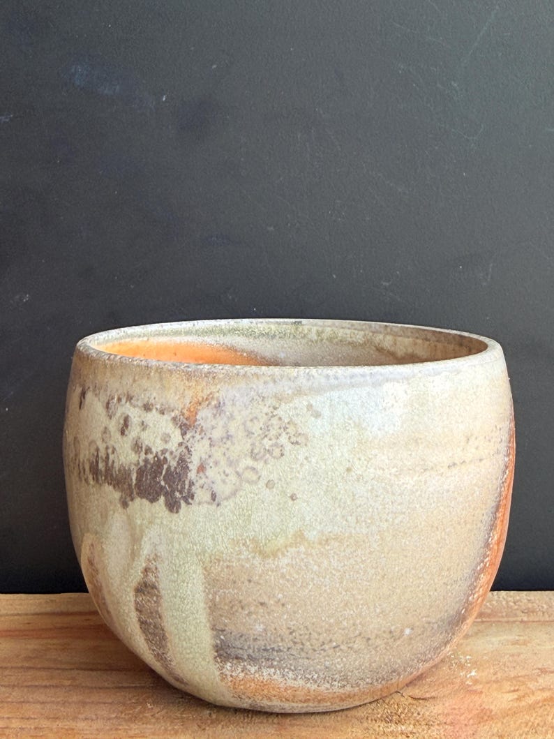 May include: A handcrafted ceramic bowl with a rounded shape. The bowl features a speckled, textured surface with a blend of beige, brown, and orange hues. The interior has an orange glaze. The bowl sits on a wooden surface.