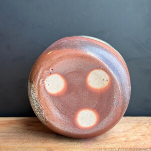 May include: A handmade ceramic vessel with a reddish-brown exterior and three white circular accents on the bottom. The vessel sits on a wooden surface against a dark background. The pottery has a rustic, textured appearance.
