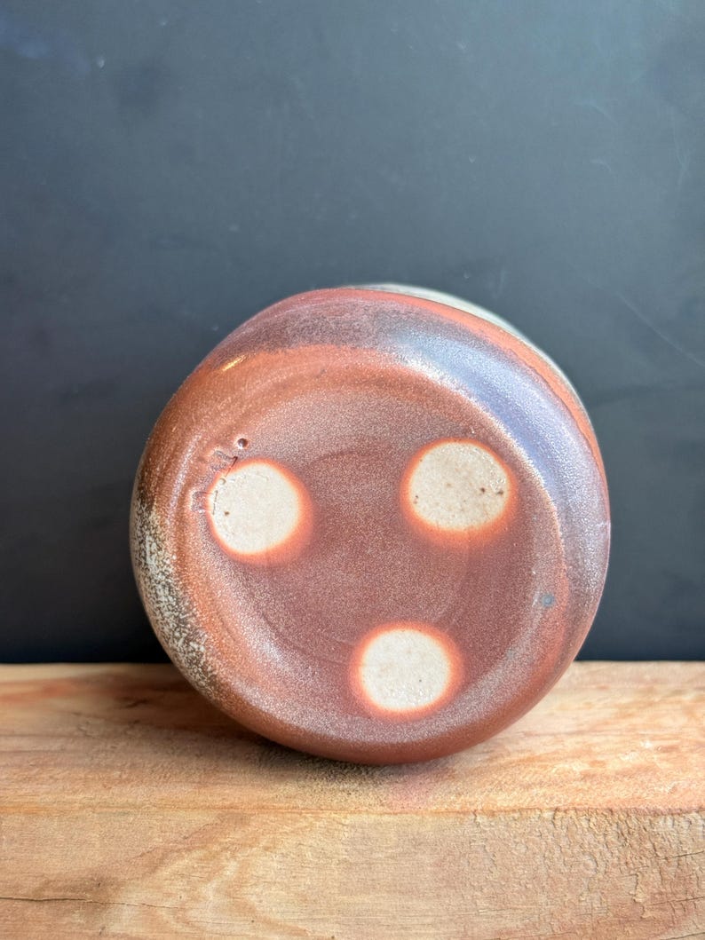 May include: A handmade ceramic vessel with a reddish-brown exterior and three white circular accents on the bottom. The vessel sits on a wooden surface against a dark background. The pottery has a rustic, textured appearance.