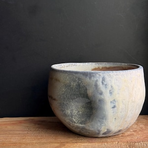 May include: A handcrafted ceramic bowl with a rounded shape and a wide opening. The bowl features a mottled, textured surface with shades of gray, blue, and cream. It sits on a wooden surface against a dark background, showcasing its unique design.