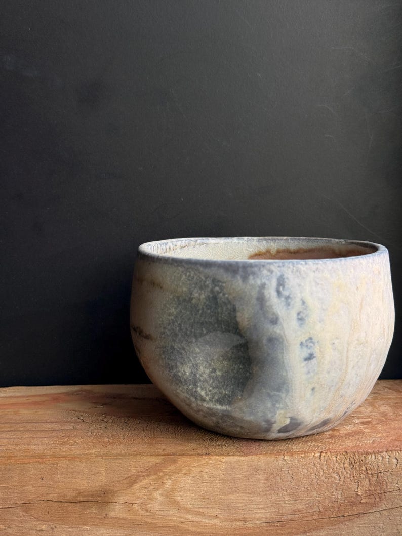 May include: A handcrafted ceramic bowl with a rounded shape and a wide opening. The bowl features a mottled, textured surface with shades of gray, blue, and cream. It sits on a wooden surface against a dark background, showcasing its unique design.