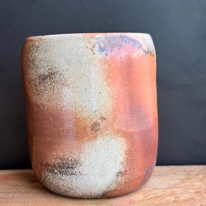 May include: A handmade ceramic tumbler with a rounded base and a slightly flared top. The exterior features a blend of colors, including white, orange, and brown, with a textured, mottled appearance. The tumbler sits on a wooden surface against a dark background.
