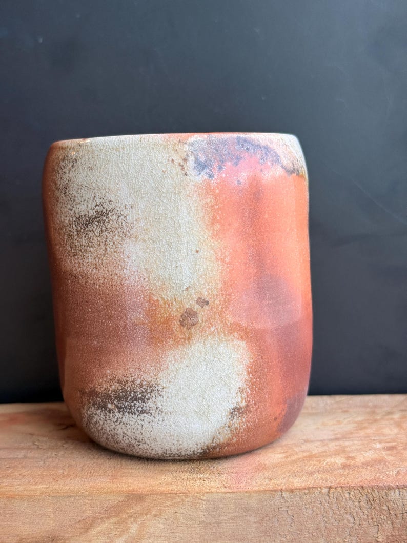 May include: A handmade ceramic tumbler with a rounded base and a slightly flared top. The exterior features a blend of colors, including white, orange, and brown, with a textured, mottled appearance. The tumbler sits on a wooden surface against a dark background.