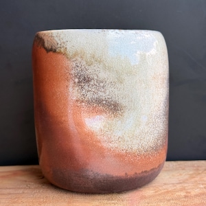 May include: A handmade ceramic tumbler with a unique glaze. The colors transition from a dark brown base to shades of orange, cream, and white at the top. The cylindrical cup has a smooth, rounded shape and is suitable for beverages.