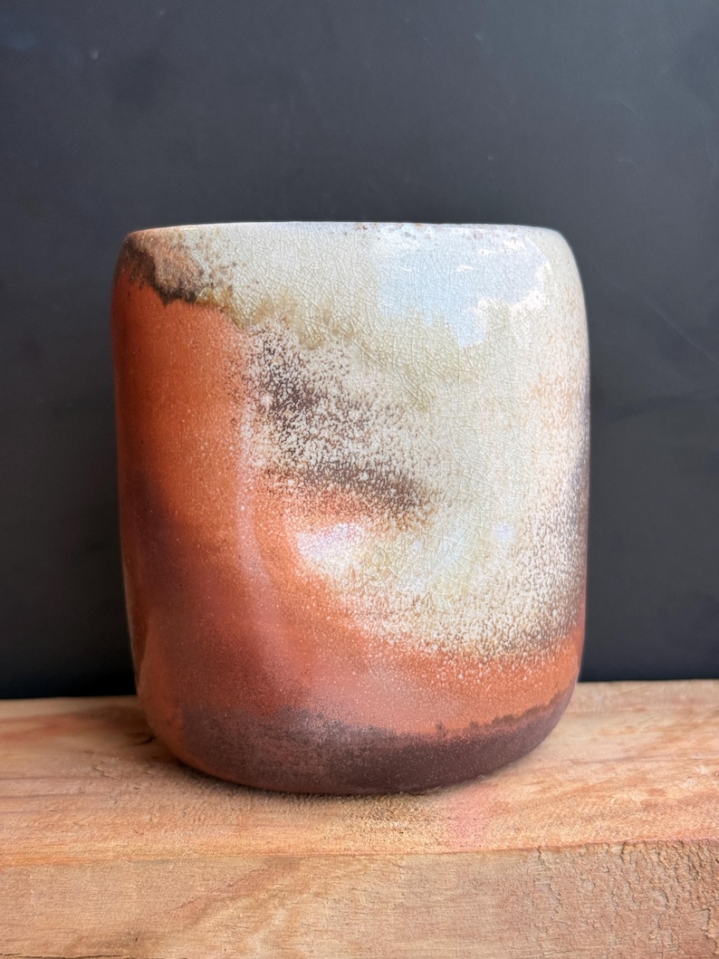May include: A handmade ceramic tumbler with a unique glaze. The colors transition from a dark brown base to shades of orange, cream, and white at the top. The cylindrical cup has a smooth, rounded shape and is suitable for beverages.