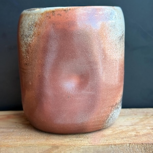 May include: A handmade ceramic mug with a rounded shape and a reddish-brown glaze. The mug has a textured surface with lighter, speckled areas near the rim. The mug sits on a wooden surface against a dark background.