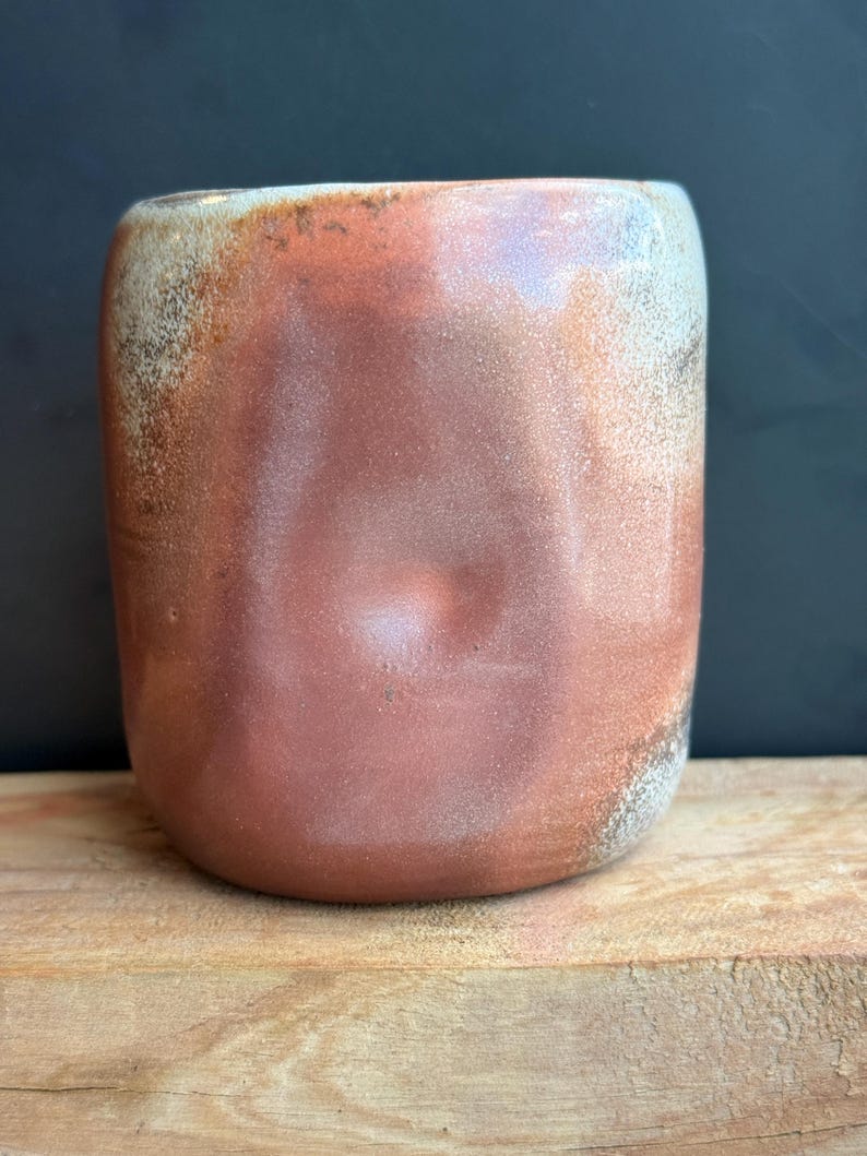 May include: A handmade ceramic mug with a rounded shape and a reddish-brown glaze. The mug has a textured surface with lighter, speckled areas near the rim. The mug sits on a wooden surface against a dark background.