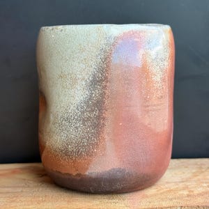 May include: A handmade ceramic mug with a unique color gradient. The mug transitions from a light beige at the top to shades of brown and reddish-orange towards the base. The mug has a rounded shape and a textured surface.