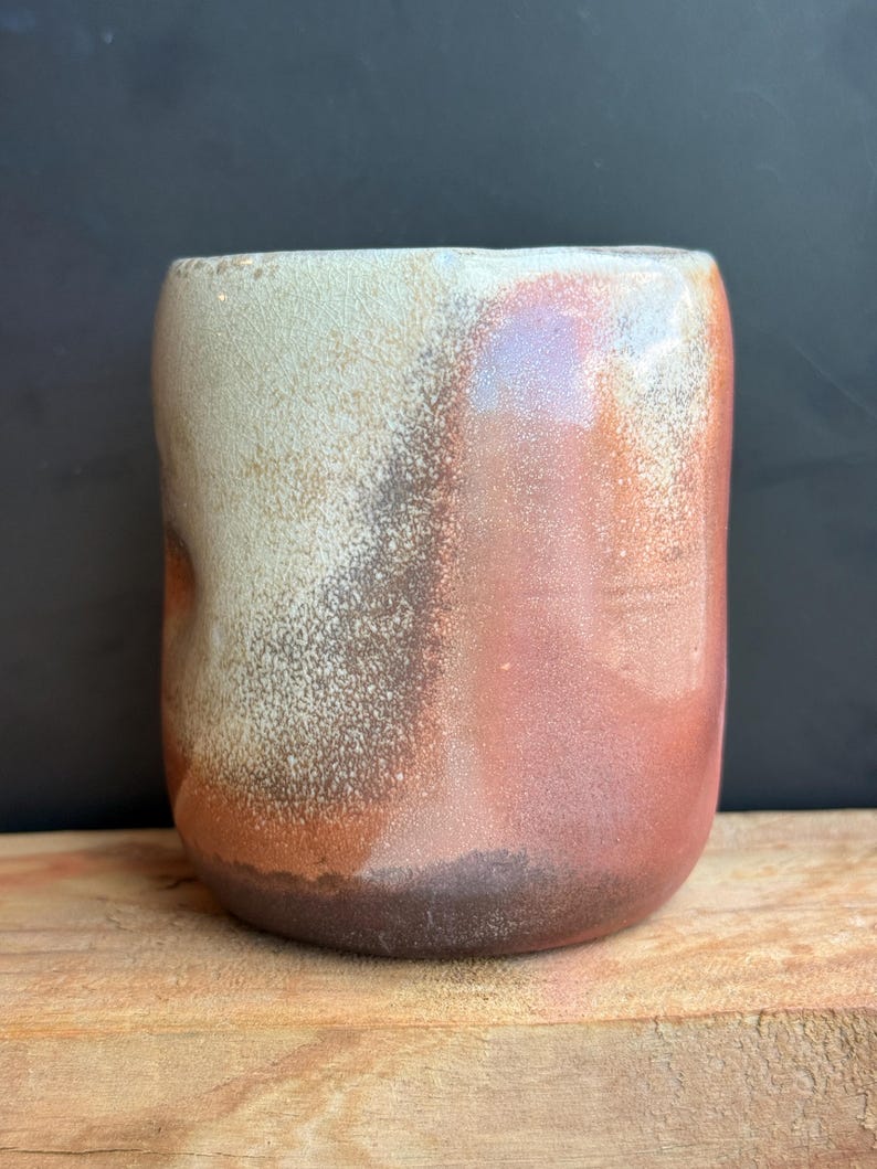 May include: A handmade ceramic mug with a unique color gradient. The mug transitions from a light beige at the top to shades of brown and reddish-orange towards the base. The mug has a rounded shape and a textured surface.