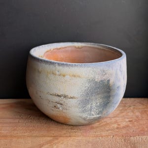 May include: A handcrafted ceramic bowl with a rounded shape. The exterior features a mottled, textured surface with shades of cream, gray, and blue. The interior has a warm, orange hue. The bowl sits on a wooden surface.