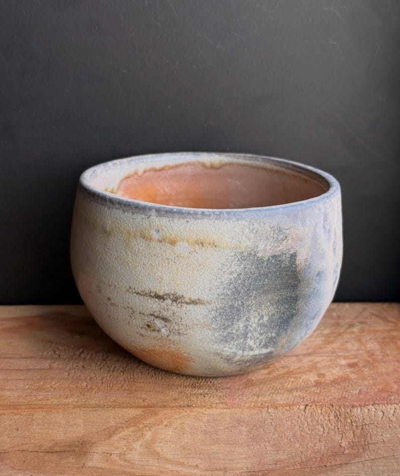 May include: A handcrafted ceramic bowl with a rounded shape. The exterior features a mottled, textured surface with shades of cream, gray, and blue. The interior has a warm, orange hue. The bowl sits on a wooden surface.