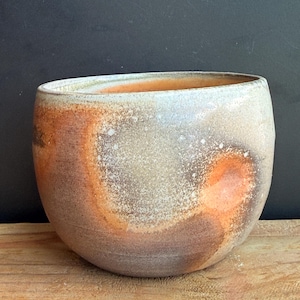 May include: A handcrafted ceramic bowl with a wide opening. The bowl features a speckled, textured surface with a blend of colors, including brown, orange, and white. The bowl is set on a wooden surface.