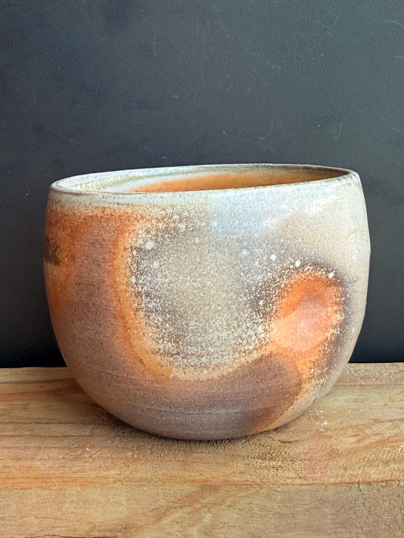 May include: A handcrafted ceramic bowl with a wide opening. The bowl features a speckled, textured surface with a blend of colors, including brown, orange, and white. The bowl is set on a wooden surface.