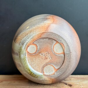 May include: A handmade ceramic vase with a rounded shape, featuring a blend of earthy tones including brown, orange, and green. The base has three circular indentations and a mountain-like symbol. The vase sits on a wooden surface.