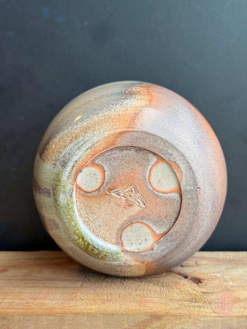 May include: A handmade ceramic vase with a rounded shape, featuring a blend of earthy tones including brown, orange, and green. The base has three circular indentations and a mountain-like symbol. The vase sits on a wooden surface.