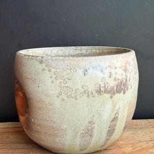 May include: A handcrafted, ceramic bowl with a wide opening and rounded shape. The bowl features a speckled, off-white glaze with brown and tan accents. The bowl is displayed on a wooden surface against a dark background.