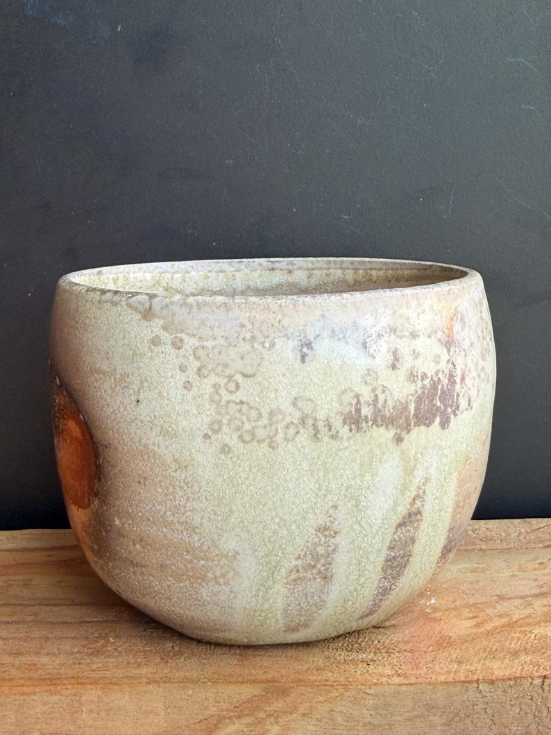 May include: A handcrafted, ceramic bowl with a wide opening and rounded shape. The bowl features a speckled, off-white glaze with brown and tan accents. The bowl is displayed on a wooden surface against a dark background.