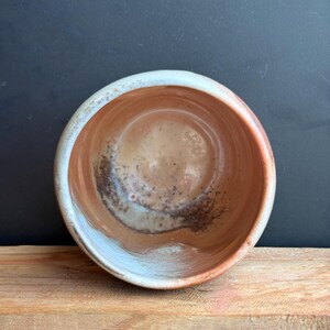 May include: A handmade ceramic bowl with a round shape. The interior features a blend of brown and tan hues with dark speckles, while the rim has a light blue and white glaze. The bowl sits on a wooden surface.