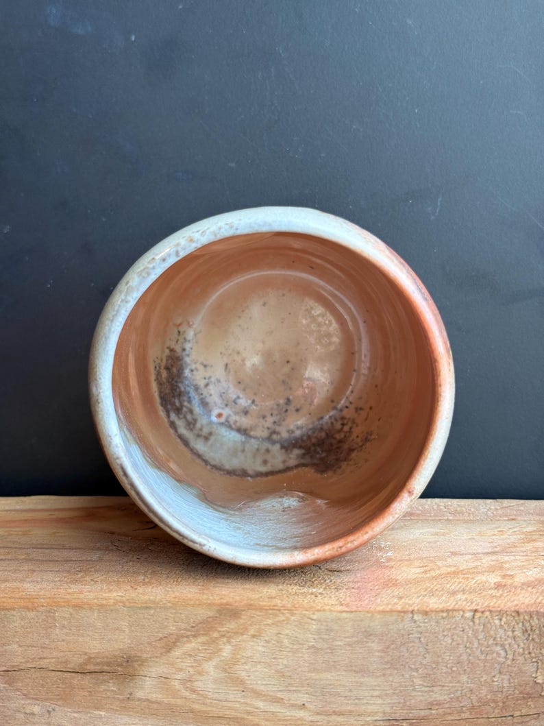May include: A handmade ceramic bowl with a round shape. The interior features a blend of brown and tan hues with dark speckles, while the rim has a light blue and white glaze. The bowl sits on a wooden surface.