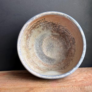 May include: A handmade ceramic bowl with a light blue rim and a textured interior. The bowl's interior features a blend of brown, gray, and white hues, creating a rustic aesthetic. The bowl is placed on a wooden surface.
