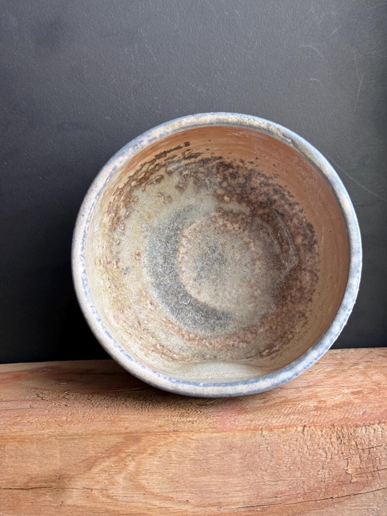 May include: A handmade ceramic bowl with a light blue rim and a textured interior. The bowl's interior features a blend of brown, gray, and white hues, creating a rustic aesthetic. The bowl is placed on a wooden surface.