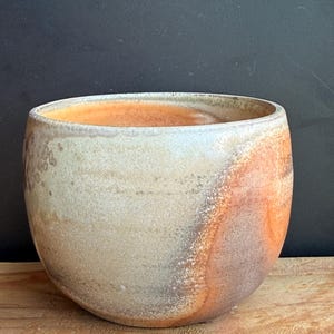 May include: A handmade ceramic bowl with a rounded shape. The bowl features a blend of colors, including beige, orange, and brown, with a textured surface. The interior has an orange hue. The bowl is sitting on a wooden surface.