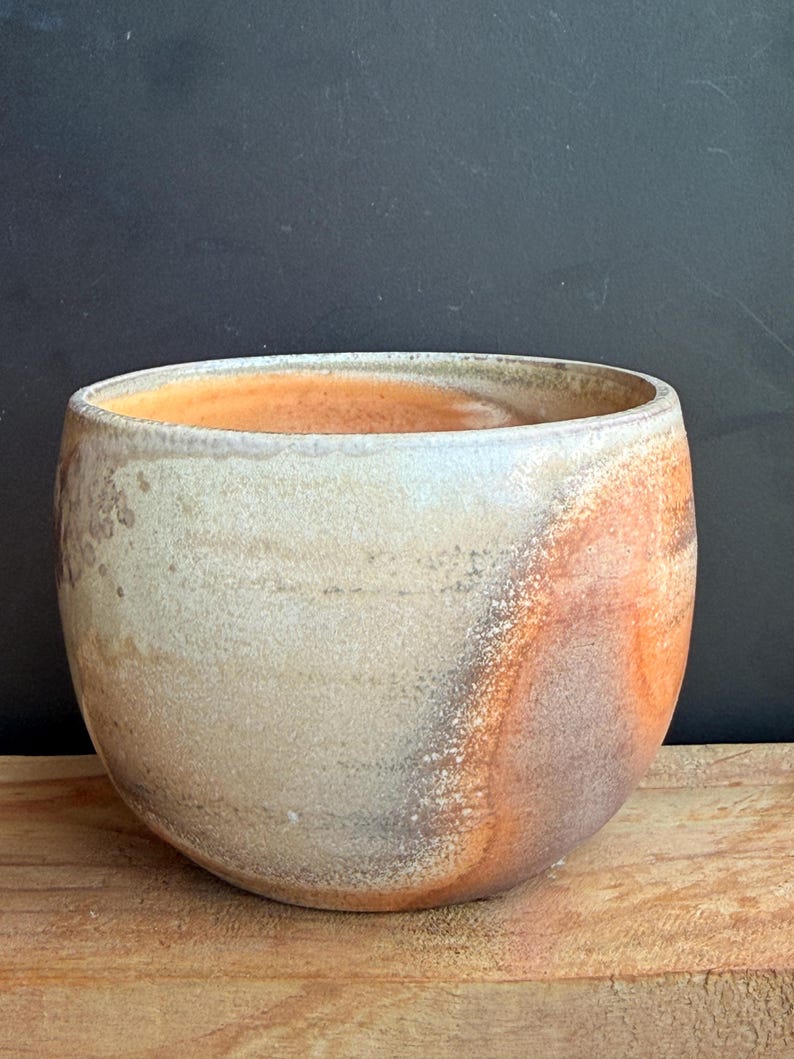 May include: A handmade ceramic bowl with a rounded shape. The bowl features a blend of colors, including beige, orange, and brown, with a textured surface. The interior has an orange hue. The bowl is sitting on a wooden surface.