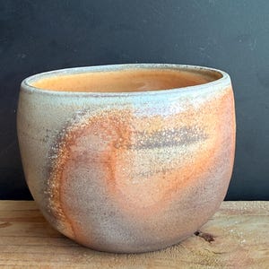 May include: A handcrafted ceramic bowl with a wide, rounded shape. The bowl features a blend of colors, including shades of orange, brown, and gray, creating a marbled effect. The interior has a warm orange hue, and the rim has a light blue tint.
