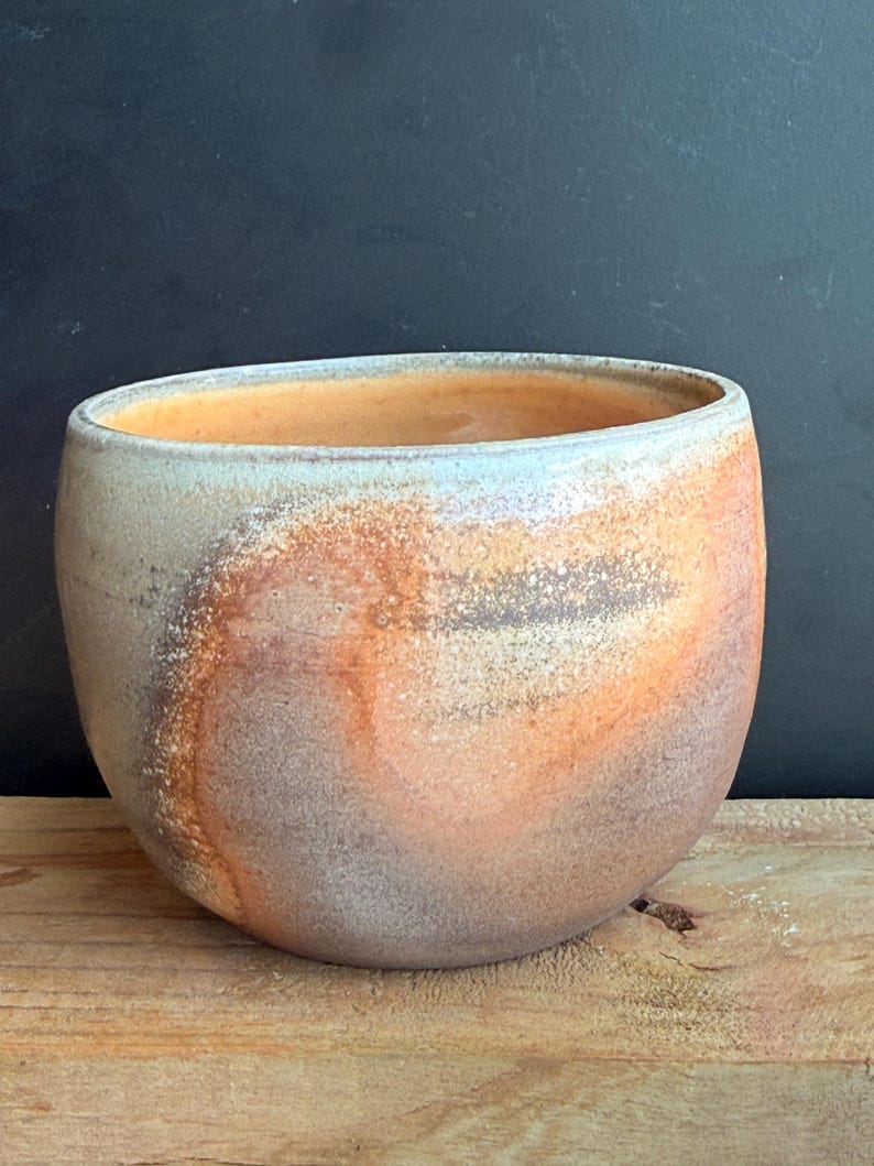 May include: A handcrafted ceramic bowl with a wide, rounded shape. The bowl features a blend of colors, including shades of orange, brown, and gray, creating a marbled effect. The interior has a warm orange hue, and the rim has a light blue tint.