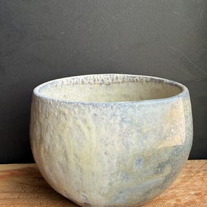 May include: A handmade ceramic bowl with a rounded shape and a textured surface. The bowl features a blend of pale yellow, blue, and gray hues, creating a unique, organic appearance. The rim has a subtle blue tint.