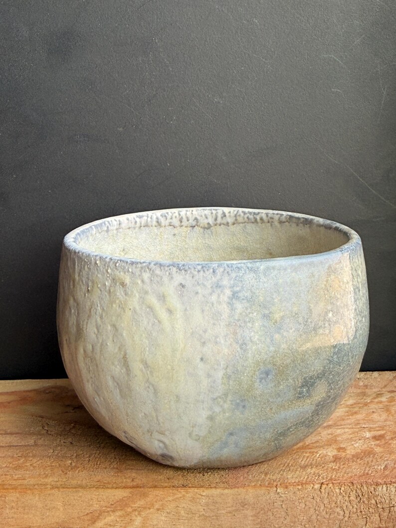 May include: A handmade ceramic bowl with a rounded shape and a textured surface. The bowl features a blend of pale yellow, blue, and gray hues, creating a unique, organic appearance. The rim has a subtle blue tint.