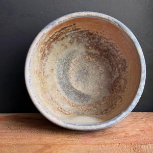 May include: A handmade ceramic bowl with a rustic, textured appearance. The bowl features a light blue rim and a blend of brown, beige, and gray tones inside. The bowl is sitting on a wooden surface.