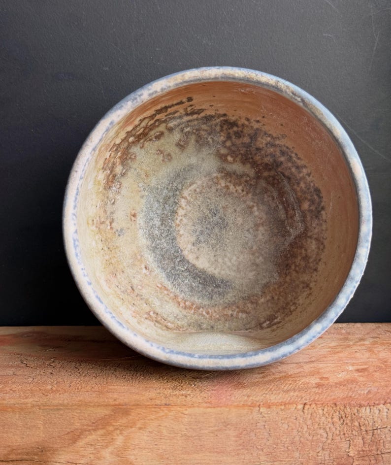 May include: A handmade ceramic bowl with a rustic, textured appearance. The bowl features a light blue rim and a blend of brown, beige, and gray tones inside. The bowl is sitting on a wooden surface.